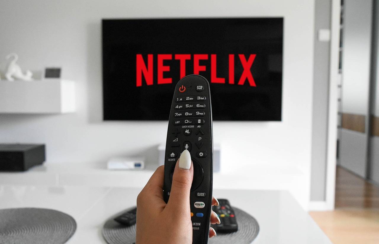 A hand holding a remote control is pointed at a TV displaying the Netflix logo in a modern living room setting.